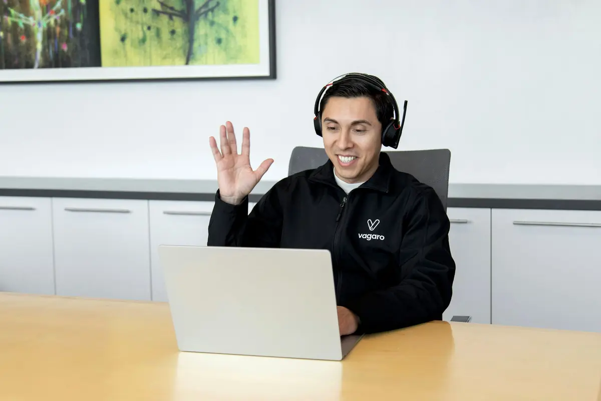 A man wearing a headset waving on a video call at his laptop.